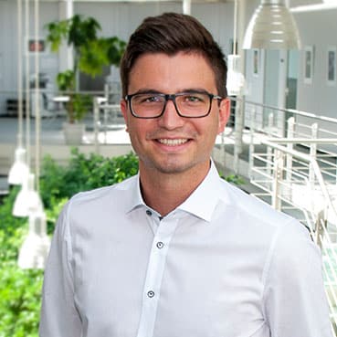 ALT-text for the picture: A happy person with glasses and blazer smiles in front of a plant, wearing a shirt with collar and sleeves.