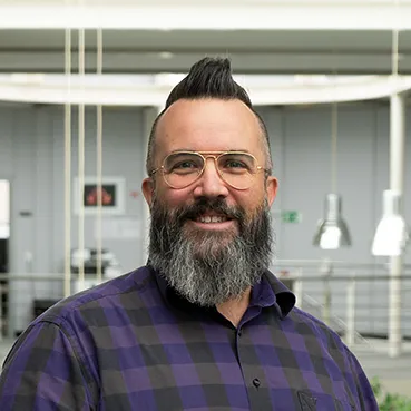 A smiling man with a beard and a visual aid, wearing a shirt with a tartan pattern and facial hair.